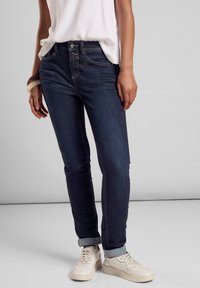 Mörkblå jeans med slim fit, uppvikta fållar och kontrasterande sömmar, i kombination med en vit texturerad topp och ljusa skor.