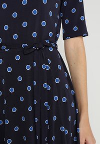 Marinblå klänning med blåa och vita prickar, korta ärmar och ett knutet midjeband. Tyget verkar mjukt med en subtil drapering.