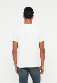 Άνδρας που στέκεται με την πλάτη προς τα έξω, φορώντα�ς ένα απλό λευκό μπλουζάκι κοντομάνικο και μπλε τζιν σε λευκό φόντο.