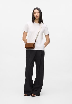 Femme portant une chemise blanche à manches courtes, un pantalon large noir, des sandales noires et un sac bandoulière marron avec une sangle beige et du texte.