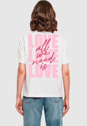 Mujer con cabello castaño hasta los hombros, vistiendo una camiseta blanca y jeans azules, la parte trasera de la camiseta dice "LOVE" en rosa y "all we need is" en letra cursiva roja.