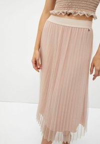 Mujer vestida con una falda midi de pliegues brillantes en color rosa claro y un top corto arrugado a juego, de pie contra un fondo blanco.