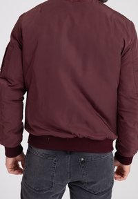 Burgundy bomberjacka med ribbad nederkant och ärmslut, med en slät yta och en enkel design. Grå jeans med bakfickorna synliga.