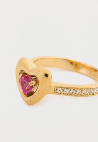 Χρυσό δαχτυλίδι με καρδιά σε σχήμα ροζ πολύτιμου λίθου στο κέντρο, με μια σειρά από διαυγείς κρυστάλλους στη ζώνη. Ορατό εγχάρακτο όνομα μάρκας.