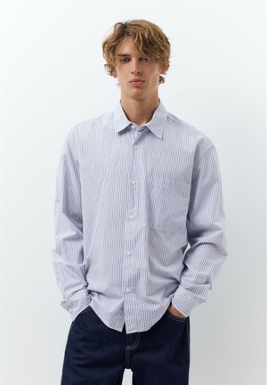 Ung mand med blondt hår iført en langærmet hvid skjorte med tynde blå striber og mørkeblå jeans, stående med hænderne i lommerne.