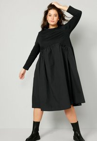 Černé šaty s dlouhým rukávem, staženým pasem, objemnou sukni a kulatým výstřihem. Tkanina vypadá hladce a strukturovaně. Černé kotníkové boty.