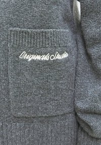 Pelēks adīts džemperis ar struktūrētu apdari, kuram ir kabata ar plāksteri un izšūts "Original Studio" logs baltā krāsā.