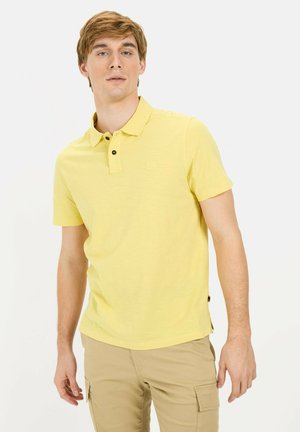 Kurzarm-Poloshirt aus leichtem gelben Baumwollstoff mit einer Knopfleiste mit zwei Knöpfen, sichtbarem Logo und strukturiertem Muster. Getragen mit khakifarbenen Hosen.