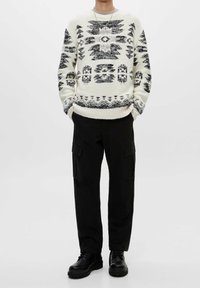 Osoba oblečená v bílém a černém vzorovaném pleteném svetru, černých pracovní kalhotách a černých šněrovacích botách, stojí s rukama v kapsách.