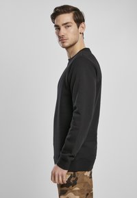 Melna džemperis ar klasisku piegriezumu, apaļu kakla izgriezumu un ribotām aprocēm. Kombinēts ar biksēm kamuflāžas rakstā.