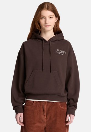 Junge Frau mit roten Haaren, die einen dunkelbraunen Kapuzenpullover mit weißem Text und rostfarbenen Cordhosen trägt.