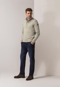Hellgrauer Pullover mit hohem Kragen, drei Knöpfen und einem blauen Patch am Ärmel, kombiniert mit dunkelblauen Jeans und braunen Schuhen.