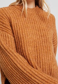 Sweter z długim rękawem w teksturowanym, prążkowanym splocie. Ciepły pomarańczowy kolor z wysokim dekoltem i lekko oversize'owym krojem. Miękki materiał.