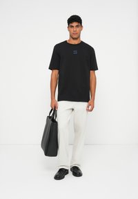 Vīrietis, kuram mugurā melna T-kreklu un baltas bikses, tur melnu audu somu, stāv uz vienkārša balta fona.