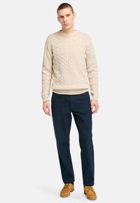 Beige Kabelstrickpullover, marineblaue Hose und sandfarbene Bootsschuhe. Der Pullover hat ein strukturiertes Design mit rundem Ausschnitt und gerippten Bündchen.