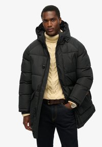 Sort polstret vinterjakke med hætte, der har lynlås og knaplukning, over en lys strikket sweater med kabelmønster, parret med mørke jeans.