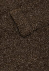 Tessuto marrone con una superficie a trama, caratterizzato da un bordo ripiegato. Il materiale sembra morbido e spesso, suggerendo calore e durevolezza.