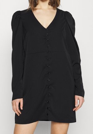 Donna che indossa un vestito nero a maniche lunghe con spalle arricciate e una fila frontale di bottoni neri dal collo all'orlo.