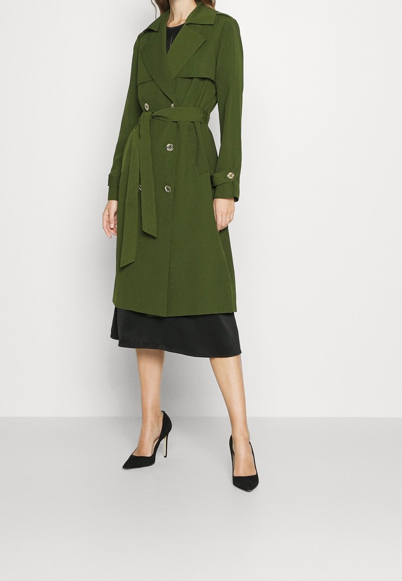 Femme portant un trench-coat vert olive ceinturé, longueur genou, sur une robe noire et des chaussures à talons hauts noires, sur un fond uni.