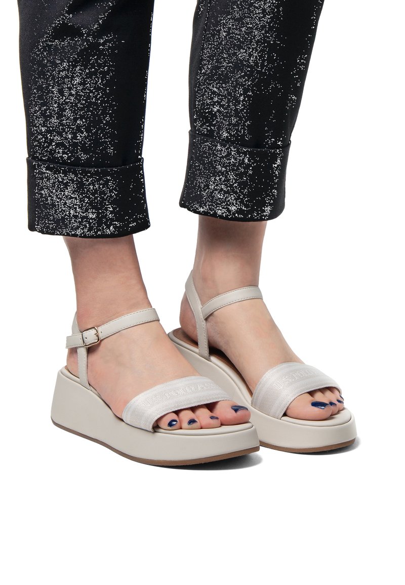 Platform sandály z světle šedé tkaniny s širokým páskem, kotníkovou přezkou a texturovanou klínovou podrážkou, doplněné černými vzorovanými kalhotami.