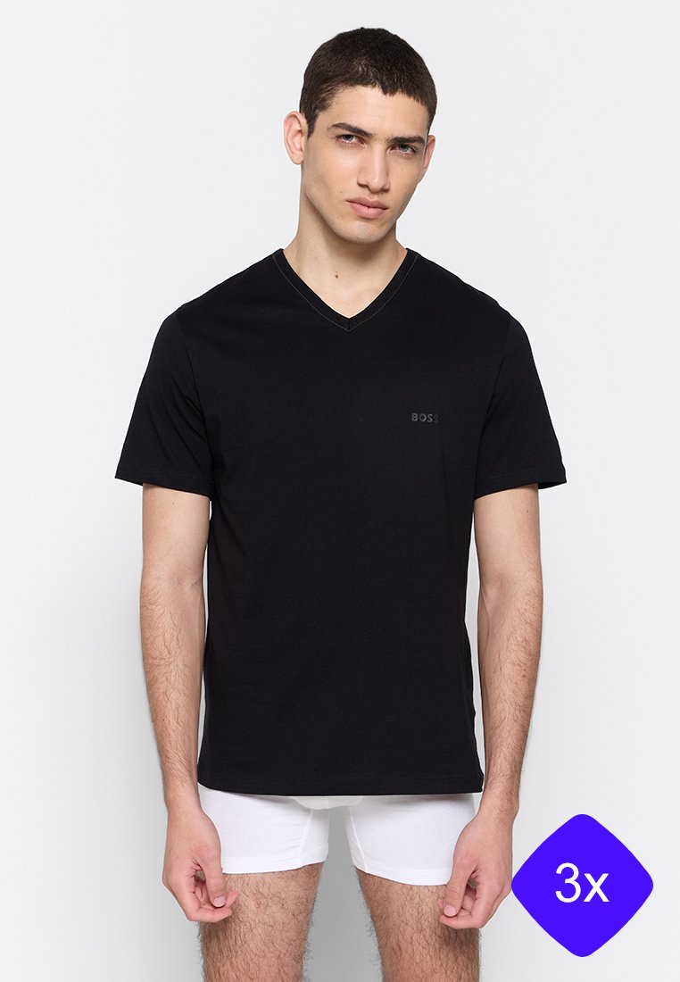 Modelo masculino vistiendo una camiseta negra de cuello en V y calzoncillos blancos, de pie frente a un fondo claro y sencillo.