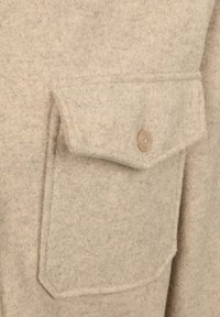 Beige wollen jas met een flapzak met een beige knoop en een subtiele geborduurde logo op de zak. Zachte, gestructureerde stof.
