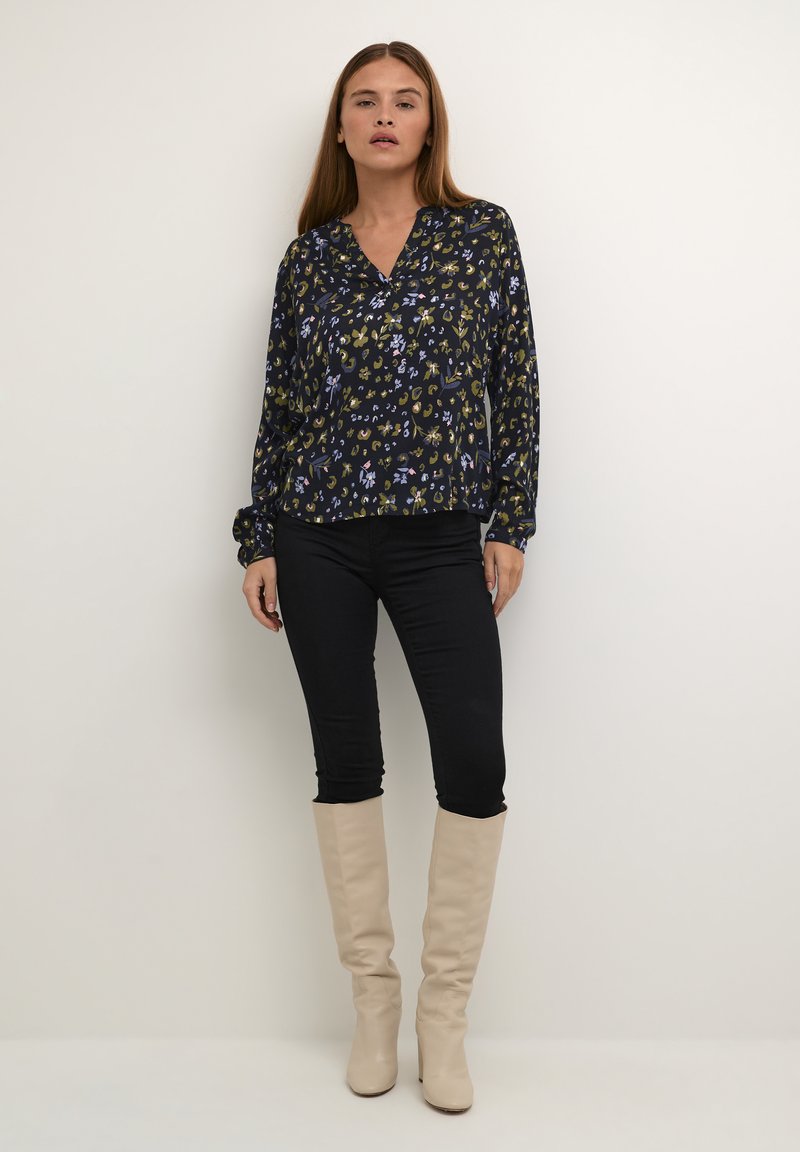Blusa a maniche lunghe con fantasia floreale nera, dotata di scollatura a V e vestibilità rilassata, abbinata a jeans skinny neri e stivali beige al ginocchio.