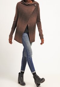 Braune, offen gestaltete Strickjacke mit Cowl-Neck und einem Knopf. Kombiniert mit blauen Jeans und schwarzen Stiefeletten. Glatte Textur, lässiges Design.