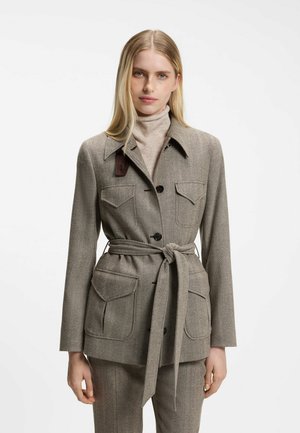 Frau trägt eine beige Jacke mit Gürtel über einem hellen Rollkragenpullover und passenden Hosen, steht vor einem schlichten weißen Hintergrund.