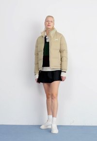 Chaqueta acolchada beige con cierre frontal, superpuesta a un suéter de punto y una camiseta polo estampada, combinada con una falda negra y zapatillas blancas.