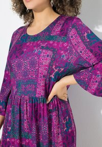 Lila Kleid mit einem Patchwork-Design, das florale und geometrische Muster in Teal und Pink zeigt. Lange Ärmel und ein geraffter Taillendetail.