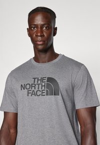 Camiseta de algodón gris con el logo de "THE NORTH FACE" impreso en negro en el pecho, cuello redondo y mangas cortas.