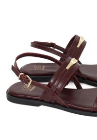 Maroons sandalen met twee dunne kruislussen, verstelbare enkelriem en gouden metalen accenten. Bovenwerk van glad leer en rubberen zool.