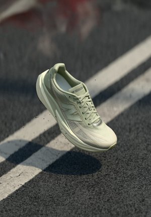 Jasnozielony but do biegania New Balance unoszący się nad nasłonecznioną asfaltową drogą z białymi malowanymi liniami.