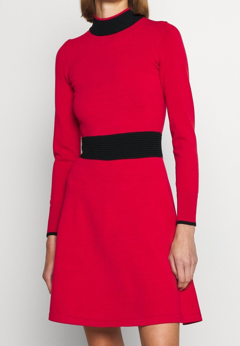 Femme portant une robe rouge à manches longues avec un col côtelé noir, une ceinture noire et des bordures noires aux poignets, debout devant un fond blanc.