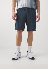 Herren Shorts in Marineblau mit Seitentaschen, aus leichtem Material, elastischem Bund und Schlitzdetails. Kombiniert mit weißen Sportschuhen und Socken.