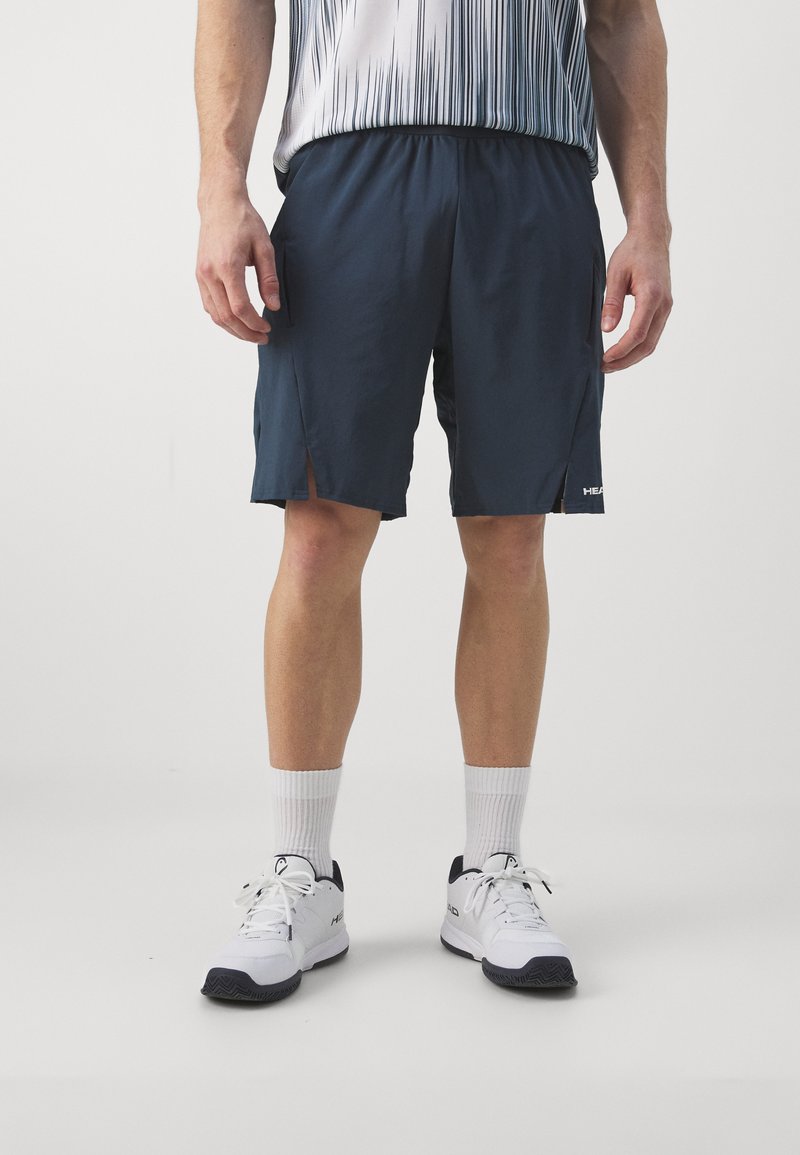 Herren Shorts in Marineblau mit Seitentaschen, aus leichtem Material, elastischem Bund und Schlitzdetails. Kombiniert mit weißen Sportschuhen und Socken.