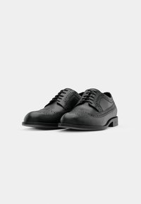 Paire de chaussures habillées brogue en cuir noir avec lacets et motifs décoratifs perforés sur un fond uni clair.
