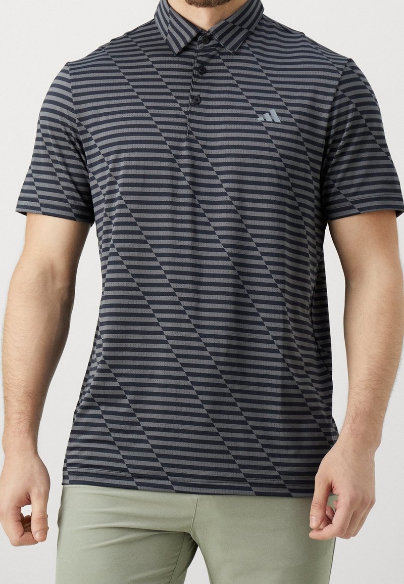 Man wearing a short-sleeve dark gray and black striped polo shirt with a collar and button placket, paired with light green pants.