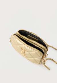 Μικρή χρυσή δερμάτινη τσάντα ώμου με ραφές σε σχέδιο πουά, δύο ανοιχτές θήκες με φερμουάρ και χρυσή αλυσίδα ως λουρί, πάνω σε λευκό φόντο.
