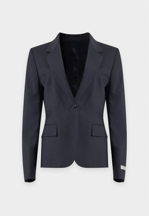 Donna che indossa un blazer scuro, top blu drappeggiato, pantaloni bianchi a gamba larga e scarpe beige in piedi su uno sfondo chiaro e uniforme.