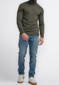 Maglione a collo alto verde scuro realizzato in un tessuto texture, abbinato a jeans blu chiaro e sneakers beige con lacci bianchi.