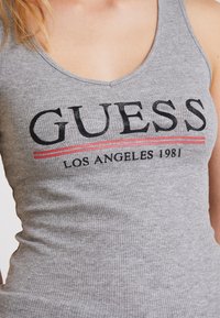 Canotta grigia a coste con grande testo nero "GUESS" con contorno rosso e nero, e "LOS ANGELES 1981" sotto. Realizzata in tessuto morbido.