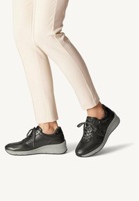 Black leather sneakers with a textured finish, featuring gray rubber soles, laces, and side zipper detail; paired with light-colored trousers.