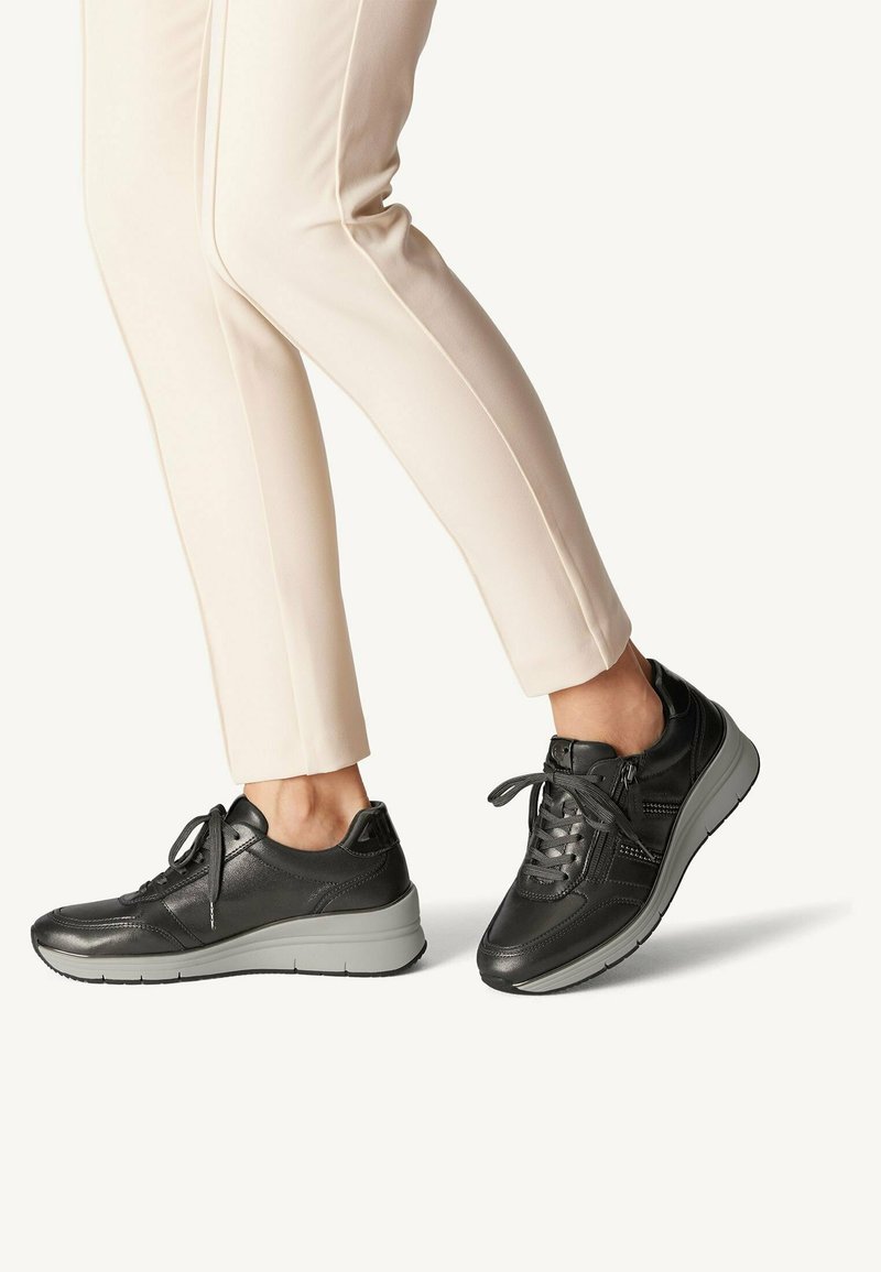 Black leather sneakers with a textured finish, featuring gray rubber soles, laces, and side zipper detail; paired with light-colored trousers.