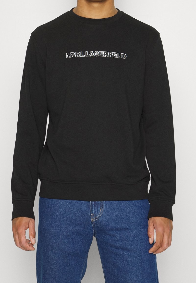Personne portant un sweat-shirt noir à manches longues avec le texte "KARL LAGERFELD" sur la poitrine et un jean bleu, debout devant un fond blanc.