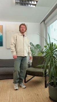 Casaco bege de botões sobre uma camisa preta, combinado com jeans largos azul claro e ténis cinzentos; ambiente interior com plantas e mobiliário.