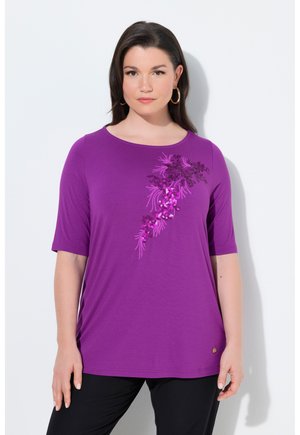 Kobieta w fioletowej bluzce z krótkim rękawem z różowym cekinowym kwiatowym wzorem z przodu, w czarnych spodniach na białym tle.