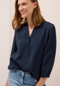 Chemise bleu marine à col en V et manches trois-quarts, dotée d'un tissu texturé et d'une coupe décontractée. Associée à un denim clair.
