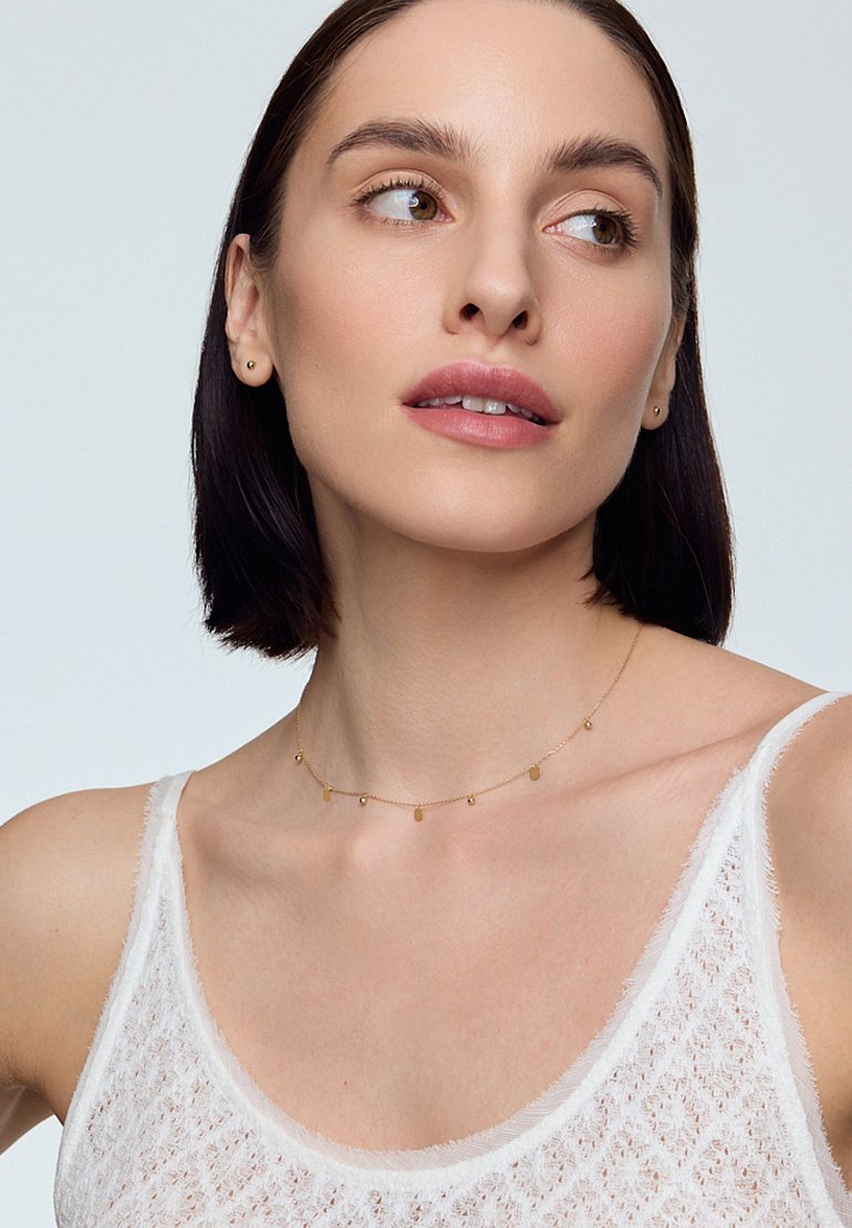 Złoty łańcuszek z małymi owalnymi i okrągłymi zawieszkami, noszony z białą, bezrękawnikową, teksturowaną bluzką na neutralnym tle.
